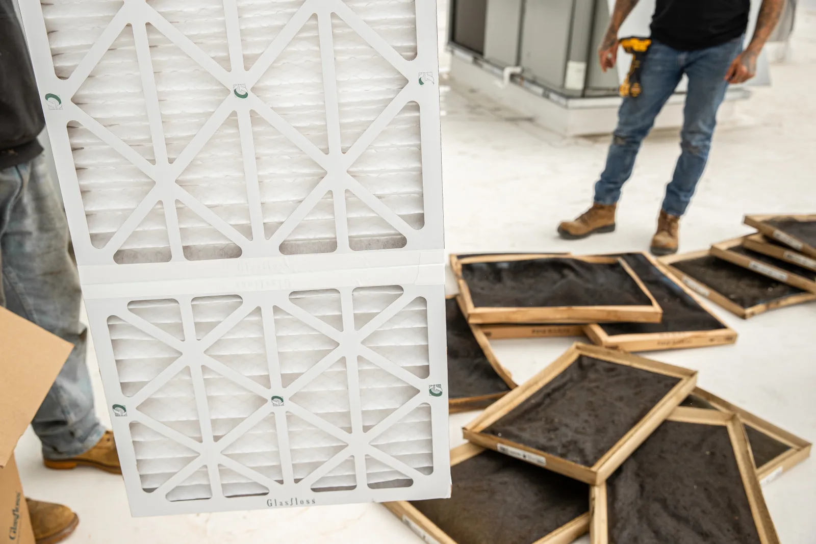 A new pleated filter held up on a commercial roof, beside a pile of clogged filters taken out of a rooftop unit
